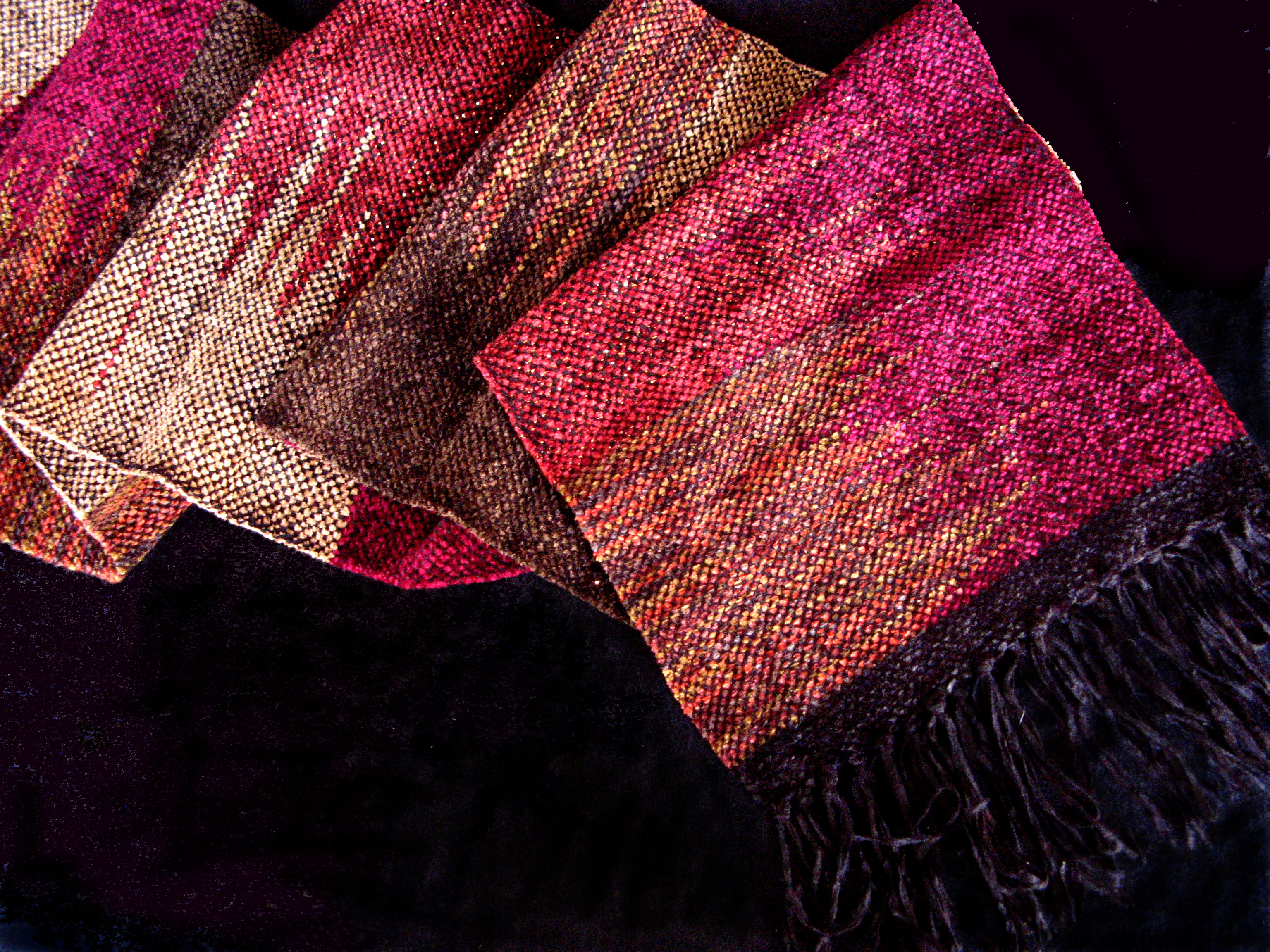 A woven scarf with three inch long black fringe lies in folds on a black background. The woven surface pattern is vertically alternating blocks of color ins hues of rose , terracotta, and brown. The blocks interlace in zigzag line that runs up the middle of the scarf.
